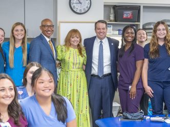 Two men in blue suits and a woman in a bright yellow patterned dress smile at the camera, surrounded by students in various colored scrubs.