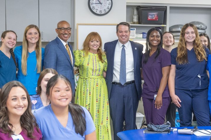 Two men in blue suits and a woman in a bright yellow patterned dress smile at the camera, surrounded by students in various colored scrubs.