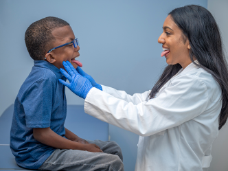 A woman wearing a doctor's lab coat stands in front of a young boy sitting on an examination table. Both are sticking their tongues out as the woman checks the boy's throat.