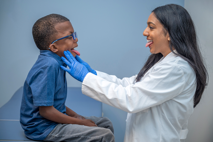A woman wearing a doctor's lab coat stands in front of a young boy sitting on an examination table. Both are sticking their tongues out as the woman checks the boy's throat.