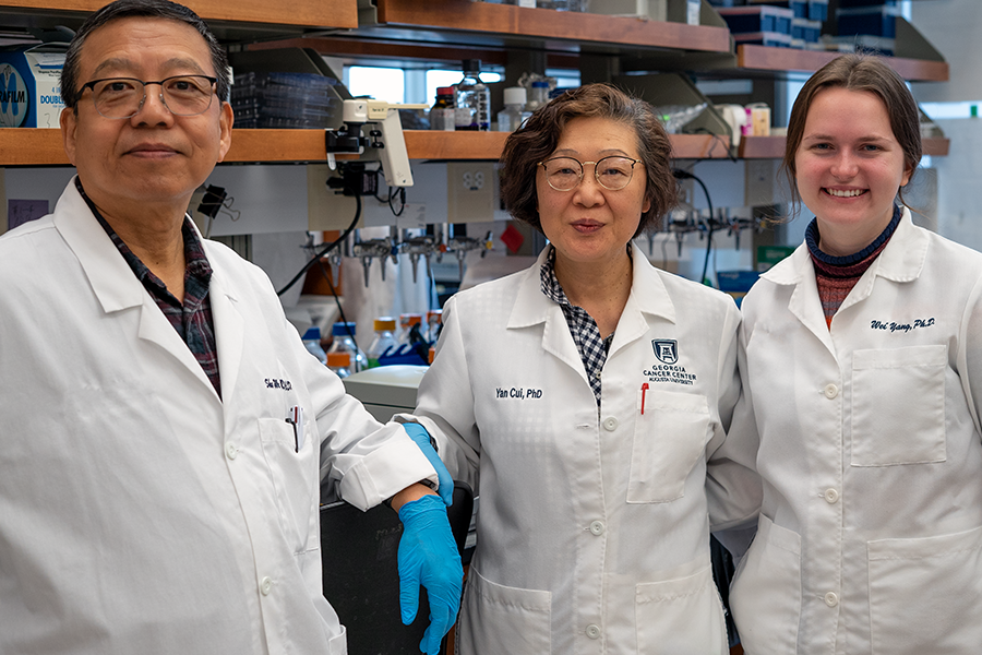 Two women and one man, all wearing scientific laboratory coats, stand in a large research lab in front of lab benches with research equipment.