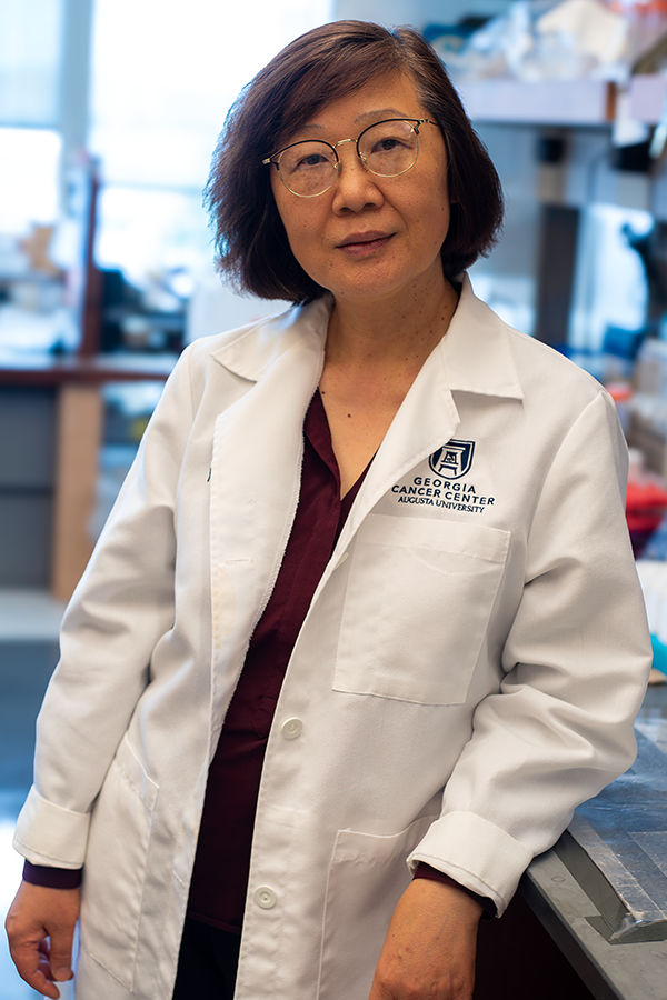 A woman in a scientific lab coat stands in a large research lab and leans against a lab bench.