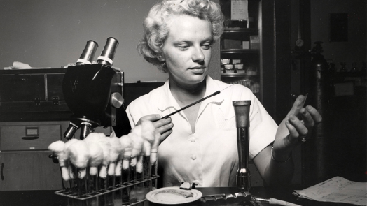 A woman sits at a laboratory bench and works with lab equipment.