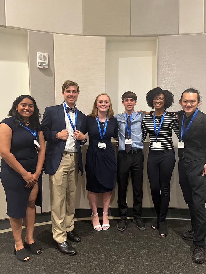 Six college students - three women and three men - stand inside a large lecture hall. All three are dressed up to give presentations.