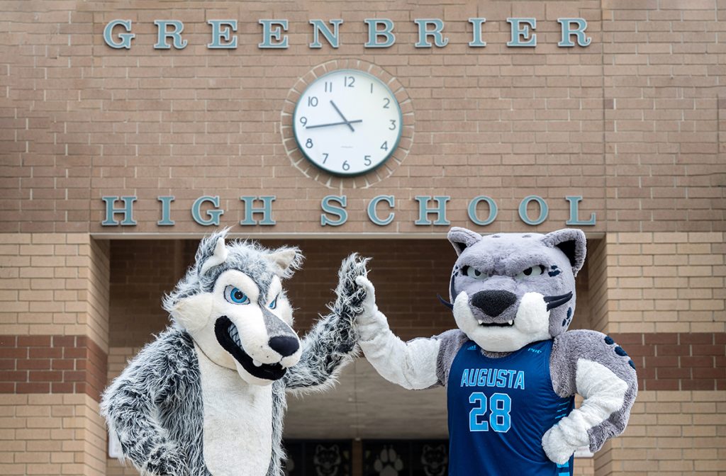 A wolf mascot high-fives the Augusta University jaguar mascot wearing a blue basketball jersey with light blue "Augusta 28" on it in front of a brick building with a clock and the words "Greenbrier High School"