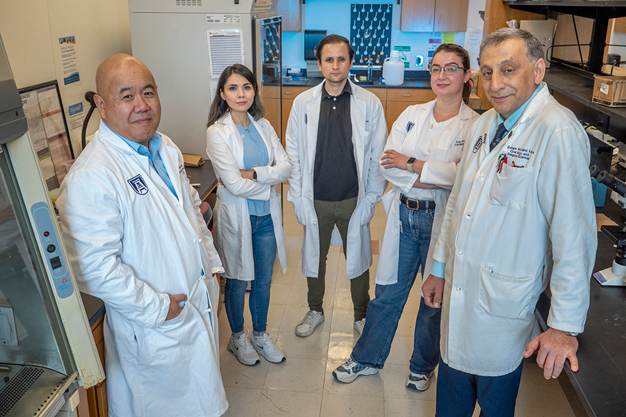 A team of five researchers, three men and two women, stand in a scientific research lab. All are wearing lab coats and are surrounded by different research tools.