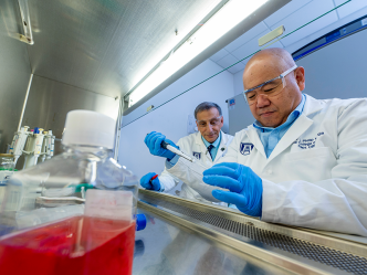 Two male scientists work on an experiment inside a lab. One is using a plastic syringe to put a liquid into a vial while the other watches from just behind.