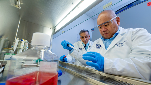 Two male scientists work on an experiment inside a lab. One is using a plastic syringe to put a liquid into a vial while the other watches from just behind.