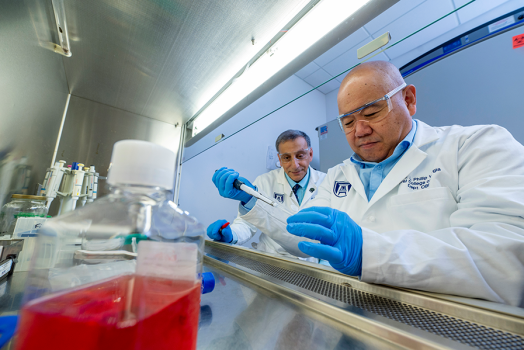 Two male scientists work on an experiment inside a lab. One is using a plastic syringe to put a liquid into a vial while the other watches from just behind.