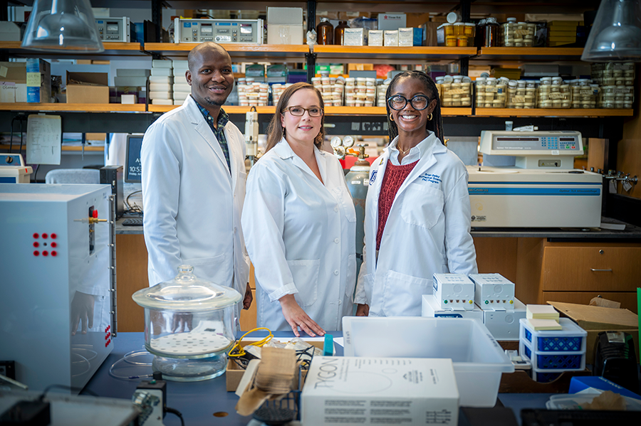 Three PhD students wearing scientific lab coats stand in a lab surrounded by various pieces of equipment.