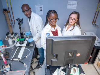 Three PhD students wearing scientific lab coats use a microscope connected to a screen.