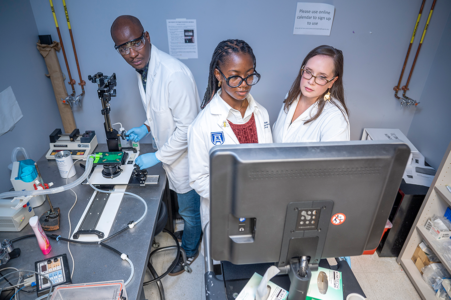 Three PhD students wearing scientific lab coats use a microscope connected to a screen.