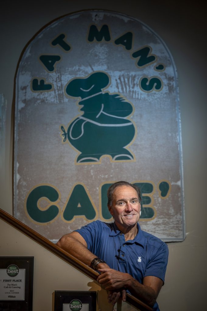 Owner of Fat Man's Cafe stands on a stairwell posing for a photo with the logo of Fat Man's Cafe on the wall behind him.