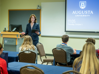 A woman stands in front of a classroom full of high school students and talks about being an educator. There is a logo for Augusta University projected onto a wall behind her.