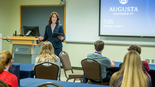 A woman stands in front of a classroom full of high school students and talks about being an educator. There is a logo for Augusta University projected onto a wall behind her.