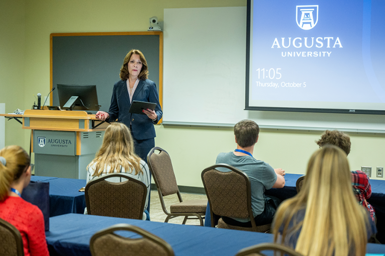 A woman stands in front of a classroom full of high school students and talks about being an educator. There is a logo for Augusta University projected onto a wall behind her.