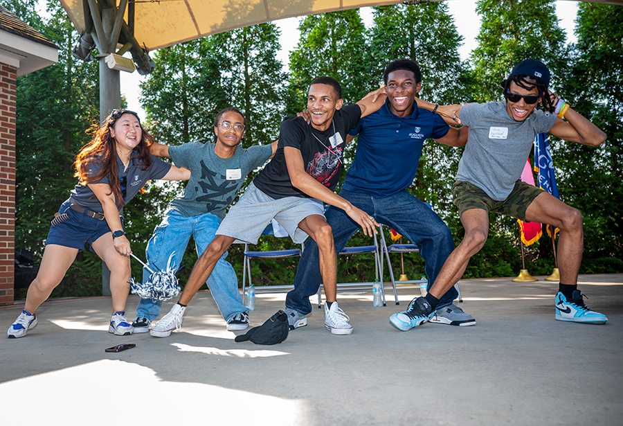 Five college students, four men and one woman, have their arms wrapped around each other's shoulders as they sway side to side.