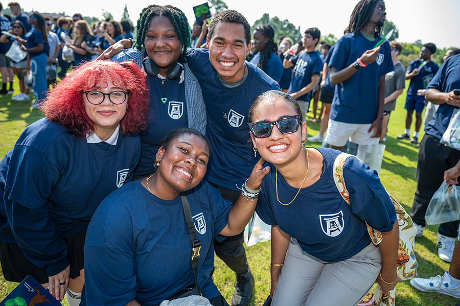 Five college students wearing shirts with a logo for Augusta University lean in toward each other to pose for a photo.