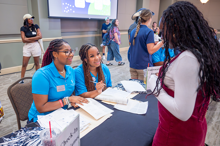 A female college freshman speaks with two female college upperclassmen at a table inside a large ballroom.