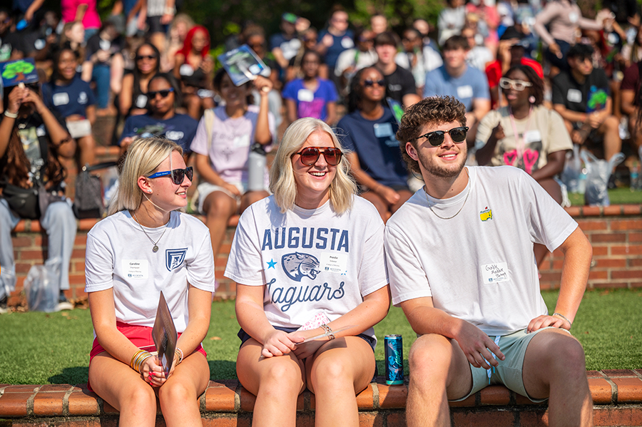 College freshmen sit around an outdoor amphitheater. Some are wearing sunglasses and others are using cardboard fans that have an image of a tree on them.