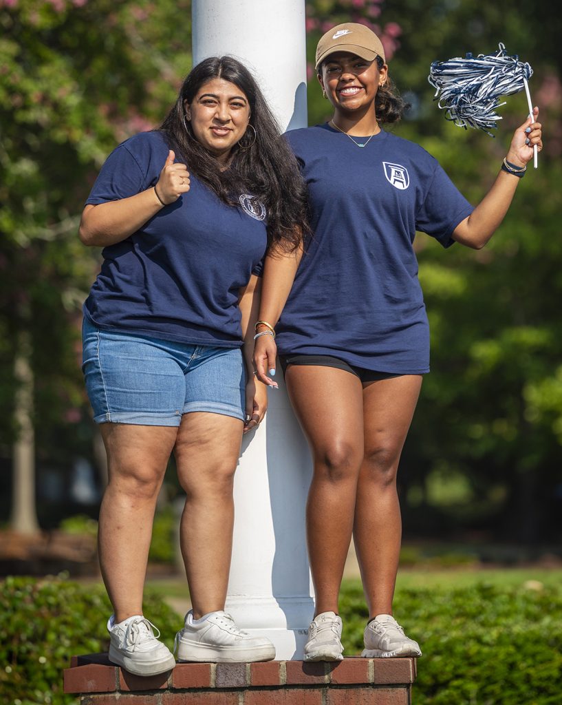 two female students wearing navy blue tshirts with the Augusta University logo in the right-hand corner stand on the base of a light pole with the woman on the right waving a pom pom