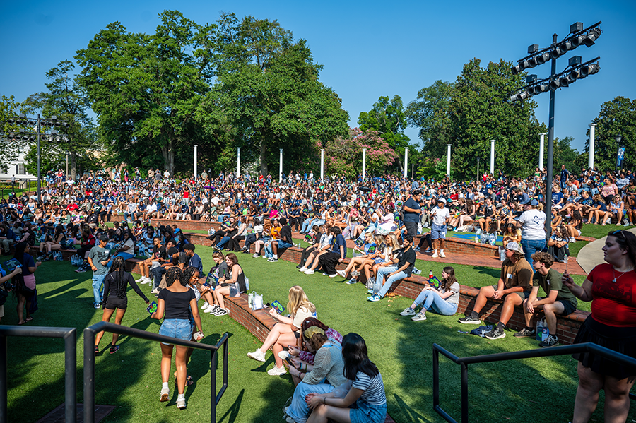 A large gathering of over 1,000 college freshmen sit and stand around an outdoor amphitheater.