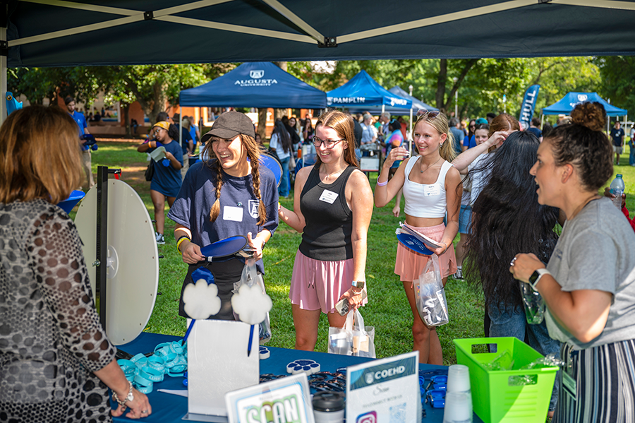 Freshman college students talk to college staff at a table in a large green space outside.