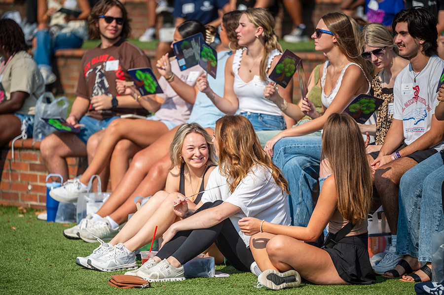 College freshmen sit around an outdoor amphitheater. Some are wearing sunglasses and others are using cardboard fans that have an image of a tree on them.