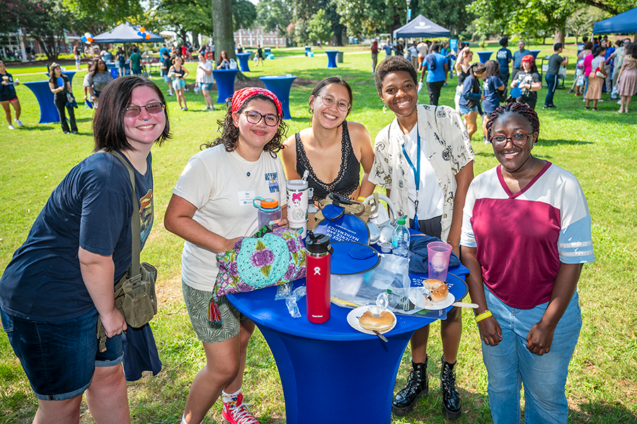 Five college freshmen women stand around a round table outside in a large greenspace with lots of people gathered in small groups around the field.