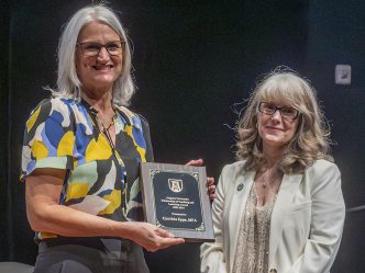 Two women stand next to each other smiling at the camera. The woman on the left is holding a vertical award.