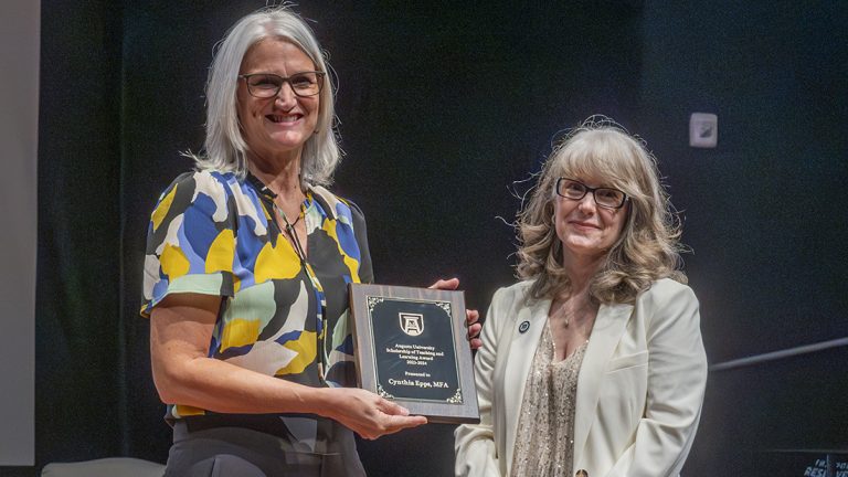 Two women stand next to each other smiling at the camera. The woman on the left is holding a vertical award.