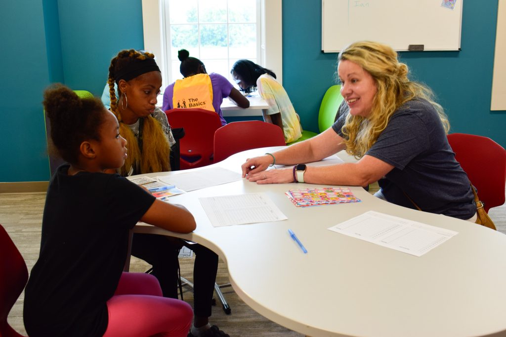 A woman leans over a table and points while smiling at two girls. She is using stickers to show them different activities.