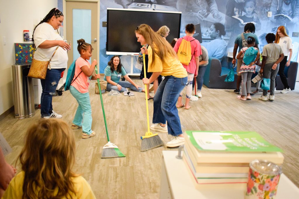 An elementary school-aged girl uses a broom to play a game with a college-aged woman inside a large space during a community event.