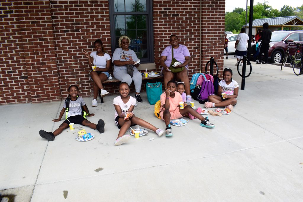 A large family sits outside on a bench and along a sidewalk, eating hotdogs and chips while enjoying a community event.