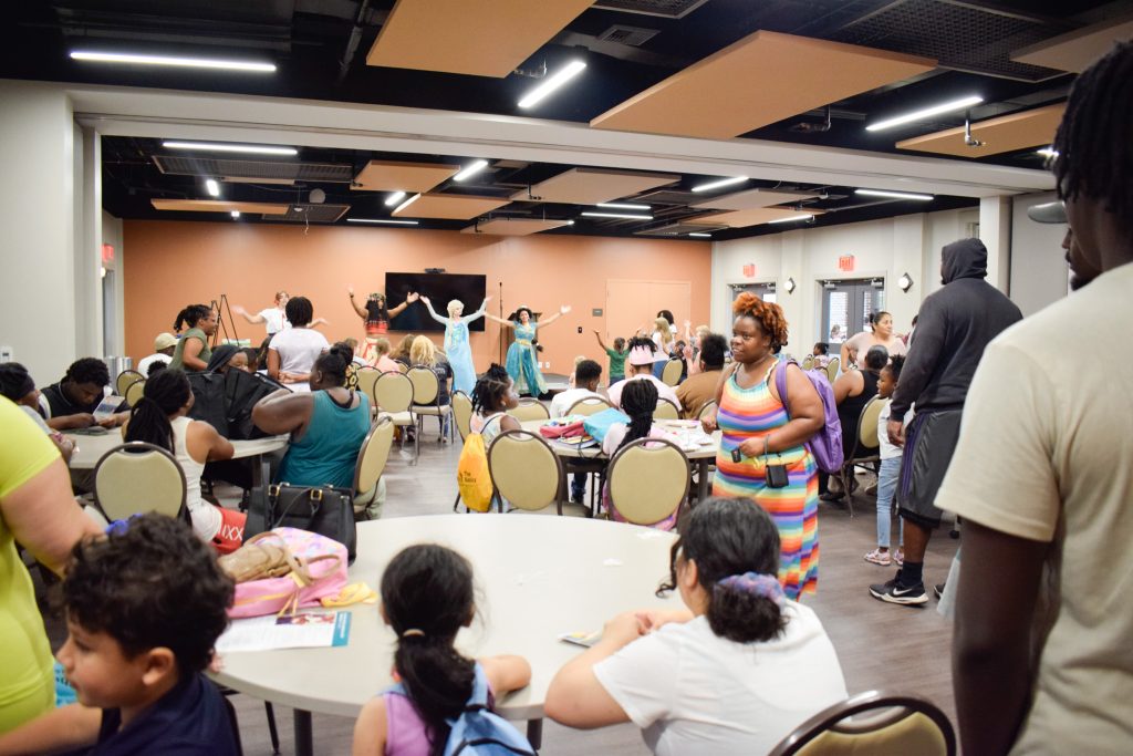 A large crowd of people sit at round tables in a large gathering space while actresses dressed up as Disney princesses dance and entertain the children in the crowd.