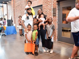 A family, including a mom, dad and five children ranging in age from a baby up to 10-years-old, stand in a large hallway surrounded by lots of people. The kids are holding bags filled with school supplies and books.