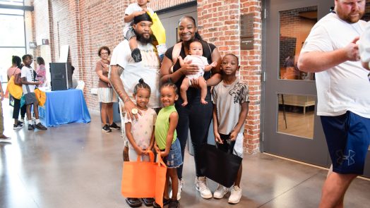 A family, including a mom, dad and five children ranging in age from a baby up to 10-years-old, stand in a large hallway surrounded by lots of people. The kids are holding bags filled with school supplies and books.