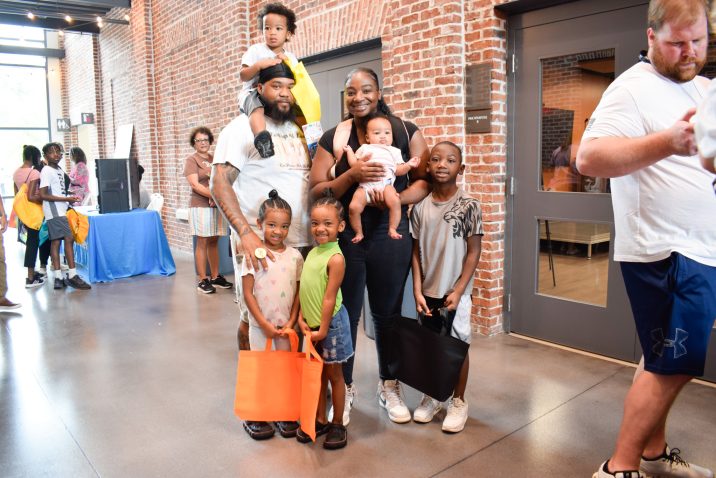 A family, including a mom, dad and five children ranging in age from a baby up to 10-years-old, stand in a large hallway surrounded by lots of people. The kids are holding bags filled with school supplies and books.