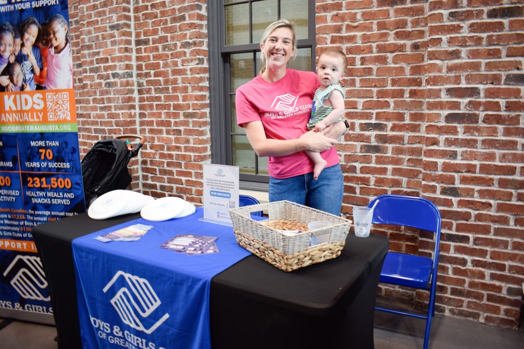 A woman holding a baby stands behind a table with signage for the Boys and Girls Club.