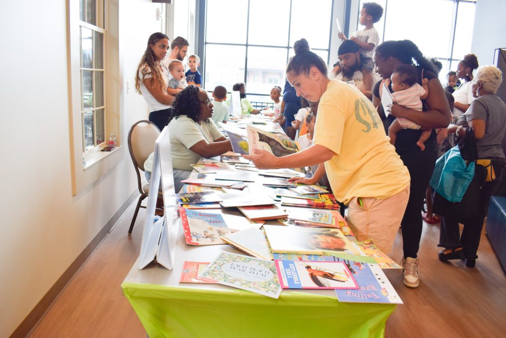 Several families gather around long tables covered in children's books. Parents look at the titles and pick out books for their children during a community event.