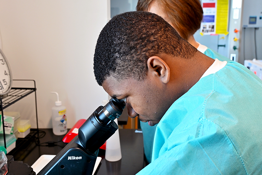 A male high school student looks into a microscope to study tumor cells while a college professor looks on.