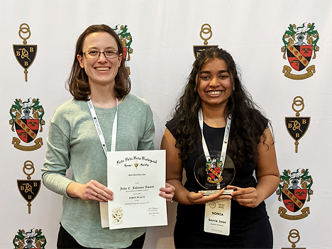 A college professor poses in front of a standing backdrop banner. She is standing next to a female college student, who has just won an award at a conference. Both hold up the certificate