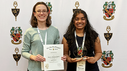 A college professor poses in front of a standing backdrop banner. She is standing next to a female college student, who has just won an award at a conference. Both hold up the certificate