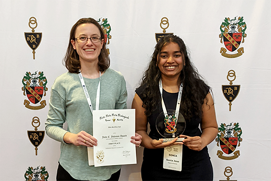 A college professor poses in front of a standing backdrop banner. She is standing next to a female college student, who has just won an award at a conference. Both hold up the certificate