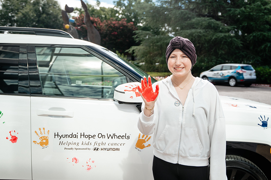 A young girl wearing a scarf around her head stands next to an SUV. her hand has paint on it from where she helped paint the car with handprints of children who have been cancer patients at Children's Hospital of Georgia.