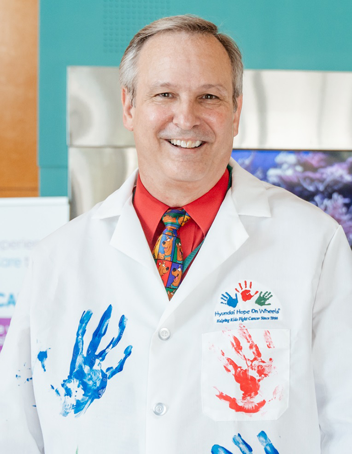 A medical doctor stands in a large atrium. His lab coat is covered in colorful handprints from children who are patients at a children's hospital.