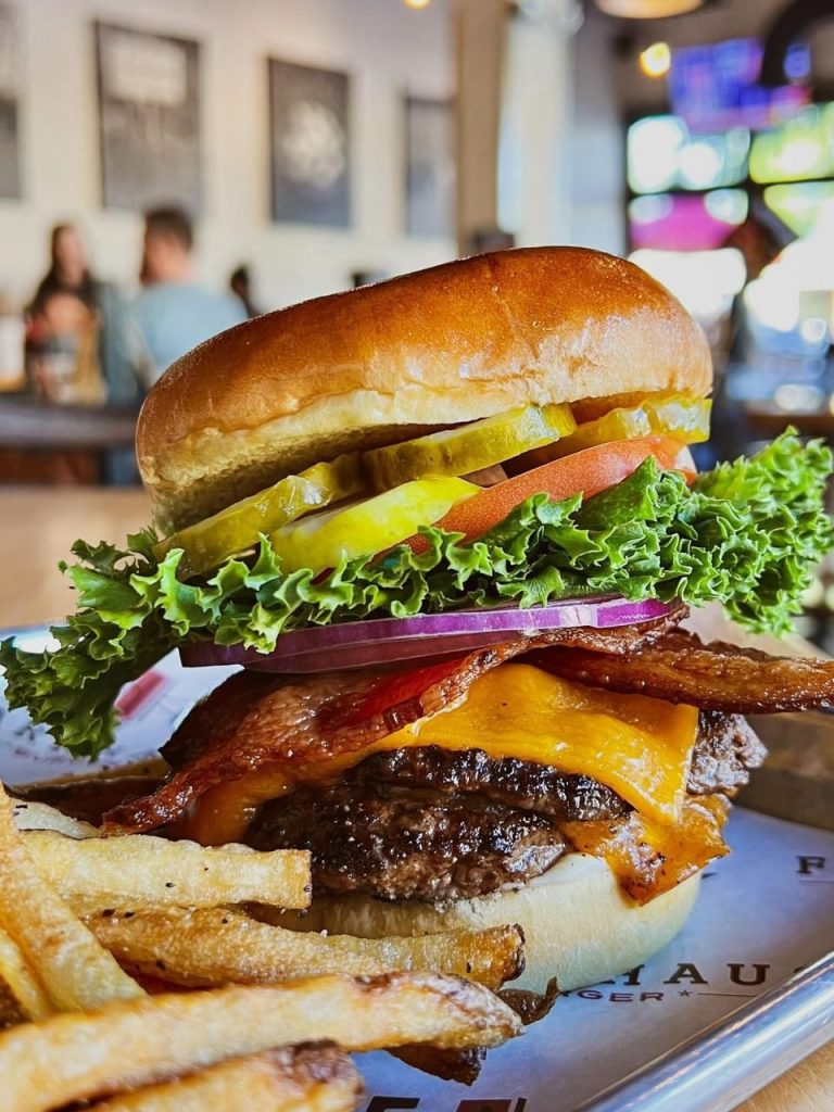 Cheeseburger with two beef patties, cheese, bacon, onion, lettuce, tomato and pickles and a side of French fries.