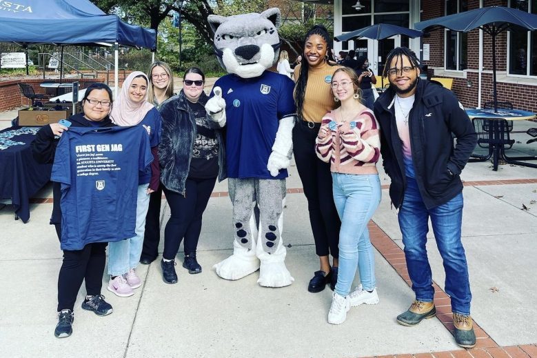 A woman and Augustus the Augusta University jaguar mascot pose for a photo outside with five female students and a male student. One of the female students holds a navy t-shirt that says "First Gen Jag."