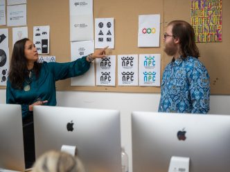 A female college professor interacts with a college student at the front of a classroom as other students look on. The professor and student are discussing logo designs that are pinned to a board between the student and professor.