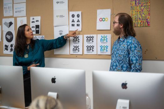 A female college professor interacts with a college student at the front of a classroom as other students look on. The professor and student are discussing logo designs that are pinned to a board between the student and professor.